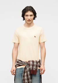 Νέος άνδρας με σκούρα μαλλιά φοράει ένα κρεμ μπλουζάκι με μικρό λογότυπο ελάφι και ένα καφέ καρό πουλόβερ δεμένο γύρω από τη μέση του.