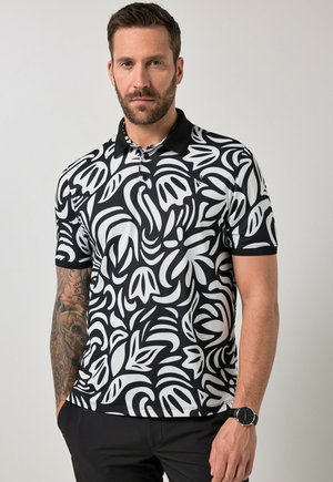 Mann mit Bart, der ein schwarz-weiß gemustertes Polohemd mit abstraktem Muster und schwarze Hose trägt, zeigt seinen tätowierten linken Arm und eine Armbanduhr am rechten Handgelenk.