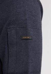 Donkerblauwe gebreide trui met een zichtbare zak en een gestikte label met de tekst "PETROL IND." in oranje. Zachte textuur, casual ontwerp.