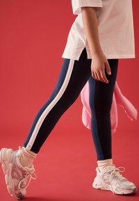 Leggings blu navy con strisce bianche ai lati, caratterizzati da una vestibilità slim. Abbinate a sneakers rosa chiaro e a un accessorio rosa appeso di lato.