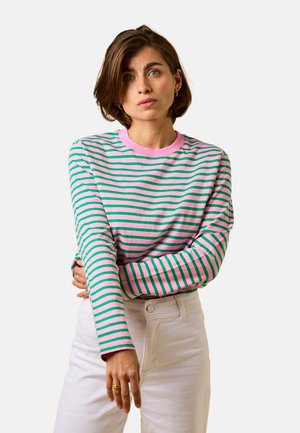 Frau mit kurzen braunen Haaren, die ein langärmeliges, rosa-grün gestreiftes Shirt und weiße Hose trägt, und den linken Ärmel mit neutralem Gesichtsausdruck richtet.