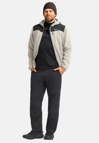 Giacca in pile con dettagli neri sulle spalle, maglietta nera a maniche lunghe, pantaloni neri e sneaker scure. Il modello indossa un berretto di maglia.