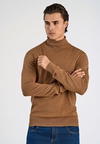 Maglione dolcevita color bronzo realizzato in tessuto morbido, con maniche lunghe e polsini a coste, abbinato a jeans blu scuro.