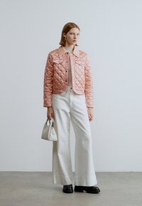 Ροζ κοντό μπουφάν με ραφές, κουμπώνει με κουμπιά ασφαλείας και έχει τσέπες, συνδυασμένο με ελαφρύ πουλόβερ, φαρδιά λευκά παντελόνια και μαύρες μπότες, κρατώντας μια μικρή λευκή τσάντα.