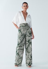 Bela srajca s kratkimi rokavi in globokim V-izrezom, v kombinaciji z visoko pasnimi zelenimi hlačami z belim vzorcem listov. Gladke teksture, sproščen kroj.