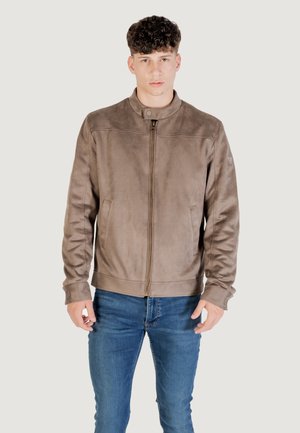 Chaqueta de cuero sintético marrón con cuello alto, cremallera frontal, bolsillos laterales y puños acanalados, llevada sobre unos vaqueros azules.