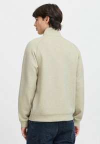 Light beige sweatshirt with a high collar, ribbed cuffs, and a relaxed fit. Smooth texture with subtle tonal pattern. Worn with dark jeans.