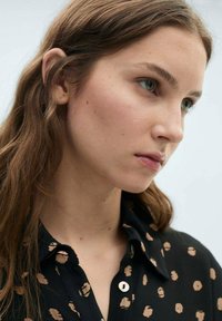 Chemise noire avec des motifs circulaires marron clair, col et boutons ; modèle avec de longs cheveux bruns ondulés et des yeux verts.