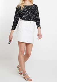 Schwarzes Polka-Dot Langarmoberteil, kombiniert mit einem weißen Jeansrock. Das Model trägt metallisch-silberne Sandalen mit Riemchen und hält eine Sonnenbrille.
