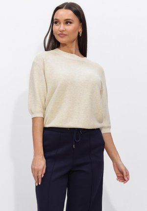 Vrouw met lang donker haar, gekleed in een gebroken wit kort mouwtruitje en marineblauwe broek met hoge taille en trekkoord, staand tegen een witte achtergrond.