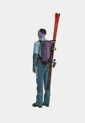 Die Skiausrüstung umfasst eine türkisfarbene und graue isolierte Jacke und Hose, einen lila Rucksack und rote Skier auf dem Rücken mit schwarzen Bindungen und Akzenten.