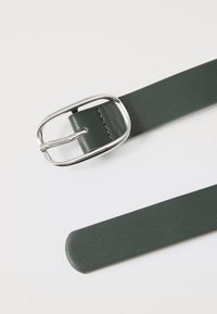 Zeleni usnjeni pas s srebrno pravokotno sponko, ki ima gladko teksturo in zaobljen konec. Preprosta detajlna šivanka.