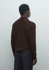 Mann in einem dunkelbraunen gerippten Pullover und einer marineblauen Hose, der steht und einer schlichten hellgrauen Wand gegenübersteht.
