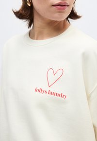 Γυναίκα που φοράει εκρού φούτερ με κόκκινη καρδιά σε περίγραμμα και ροζ κείμενο «lollys laundry» στο στήθος, και χρυσούς κρίκους σκουλαρίκια.