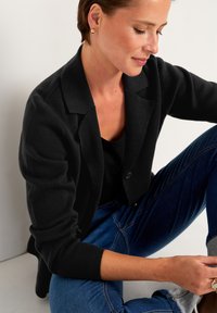 Sort strikjakke med en struktureret krave, enkelt knaplukning og optrukne ærmer, parret med en sort top og blå jeans.