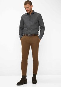 Schwarzes Hemd mit weißen Punkten zum Zumachen, braune Slim-Fit-Hose und dunkle braune Stiefeletten. Das Hemd hat einen Kragen und lange Ärmel.