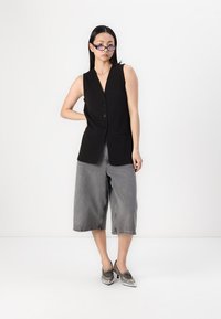 Schwarze ärmellose Weste mit Knöpfen, kombiniert mit grauen weit geschnittenen Jeansshorts und metallischen Schuhen, die ein minimalistisches Design betonen.
