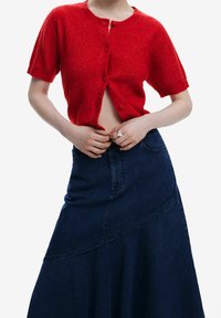 Punainen lyhythihainen neulecardigan, jossa on pyöreä pääntie ja nappikiinnitys, yhdistettynä tummansinisestä denimmistä valmistettuun maksihameeseen, jossa on epäsymmetrinen helma.