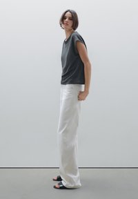 T-shirt grigio a maniche corte abbinato a pantaloni bianchi a gamba larga, con una vestibilità rilassata. Il modello indossa sandali neri a pianta piatta. Sfondo minimale.