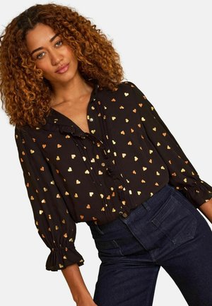 Femme aux cheveux bouclés portant un chemisier noir orné de petits cœurs dorés et un jean taille haute bleu foncé, posant devant un fond blanc.