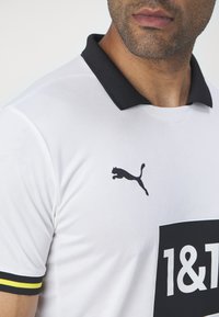 Bela športna majica z črnim ovratnikom. Ima črn logotip Puma in pravokotni črn znak s "1&1" v beli barvi. Ovratniki so v rumenih in črnih črtah.