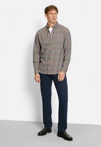 Kariertes Hemd mit roten, grünen und blauen Quadraten, mit geknöpftem Kragen, kombiniert mit dunkelblauen Hosen und schwarzen Schuhen, stehend vor neutralem Hintergrund.