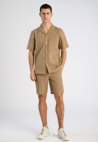 Camisa de manga corta color marrón claro y pantalones cortos a juego de tela texturizada; cuenta con dos bolsillos frontales y cierre de botón. Lleva con zapatillas blancas.