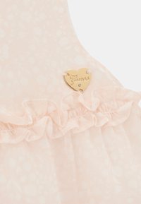 Robe rose clair avec motif floral, bordure froncée et étiquette en forme de cœur doré avec l'inscription "Guess". Texture de tissu délicat et transparent.