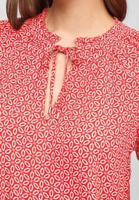 Blouse rouge à motifs avec une ouverture en forme de goutte, tissu texturé présentant des motifs floraux, et un détail de col à volants.
