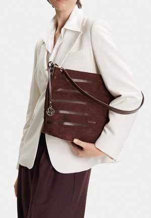 Osoba oblečená v bílém saku a tmavých kalhotách drží tmavě hnědou koženou tašku přes rameno s proužky a stříbrným přívěskem.