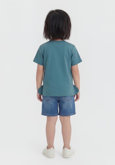 Enfant aux cheveux foncés mi-longs, debout de dos, portant un t-shirt turquoise, un short en jean et des baskets blanches sur un fond uni.