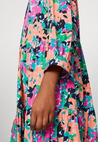 Braccio esteso verso il basso indossando un vestito lungo con maniche floreali colorate e polsini arricciati nei motivi rosa, verde, arancione, blu e nero.