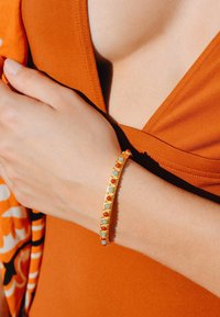 Bracelet en or avec des pierres circulaires rouges et bleu clair, présentant un bandeau lisse avec de petits accents. Porté sur une main contre un fond orange.
