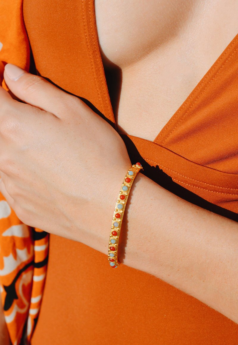 Bracelet en or avec des pierres circulaires rouges et bleu clair, présentant un bandeau lisse avec de petits accents. Porté sur une main contre un fond orange.