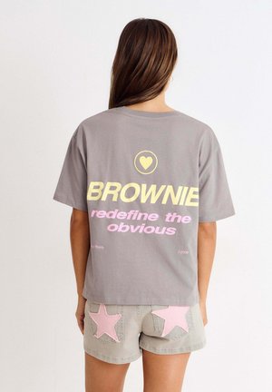 Femme aux longs cheveux bruns porte un T-shirt gris oversize avec le texte "BROWNIE redefine the obvious" et un graphique en forme de cœur, ainsi que des shorts gris avec des étoiles roses.