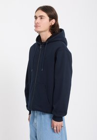Jeune homme aux cheveux mi-longs, portant un sweat à capuche zippé bleu marine et un jean bleu clair, debout et légèrement tourné vers la gauche.