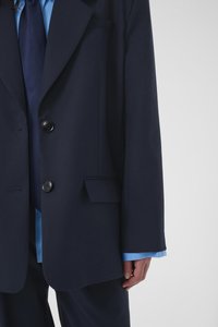 Άτομο που φοράει σκούρο μπλε σακάκι κοστουμιού πάνω από μπλε πουκάμισο με έναν ταίριαστο μπλε γραβάτα, το χέρι χαλαρό δίπλα τους μπροστά σε απλό φόντο.