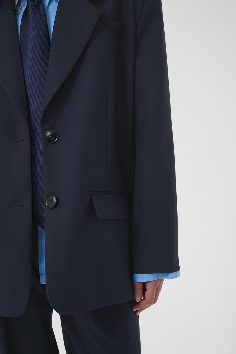 Personne portant une veste de costume bleu marine foncé sur une chemise bleue avec une cravate bleu marine assortie, main détendue le long du corps sur un fond uni.