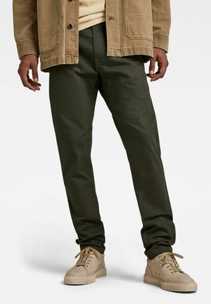 Man met olijfgroene broek, beige jas en beige sneakers, staand tegen een effen witte achtergrond.