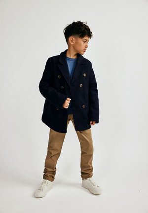 Tumesinine kahekordne jope, mis on valmistatud villasegusest, suurte nööpidega, kantud sinise särgi peal, koos beežide pükste ja valgete tossudega.