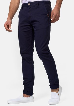 Mann trägt dunkelblaue Slim-Fit-Hose und weiße Slip-On-Sneaker, steht mit entspannt an den Seiten hängenden Händen vor weißem Hintergrund.