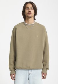 Sweatshirt vert olive avec col rond et poignets côtel�és. Présente un petit logo sur la poitrine et une coupe décontractée. Porté sur un T-shirt blanc.