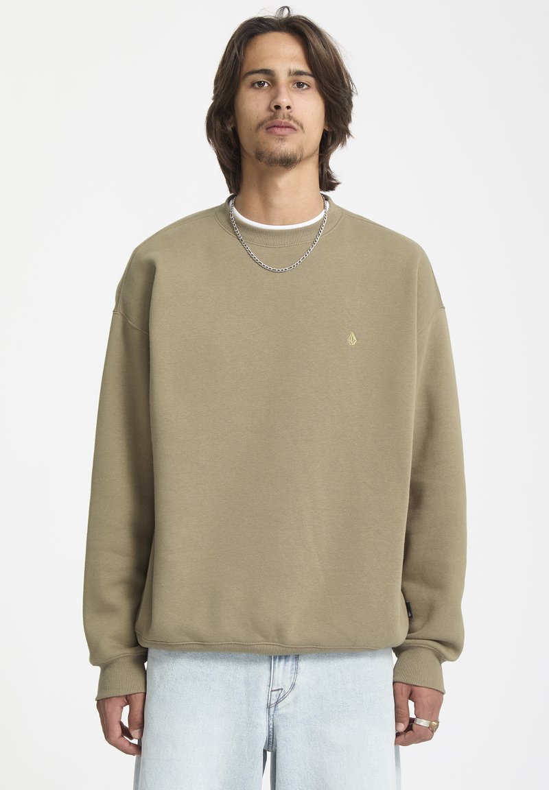Sweatshirt vert olive avec col rond et poignets côtelés. Présente un petit logo sur la poitrine et une coupe décontractée. Porté sur un T-shirt blanc.