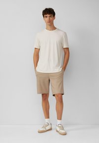 Helles beige T-Shirt mit kurzen Ärmeln, kombiniert mit sandfarbenen Shorts im entspannten Schnitt. Texturiertes Material, minimales Design ohne sichtbare Muster.