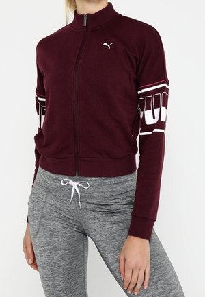 Femme portant une veste Puma bordeaux zippée avec un logo sur la poitrine et un texte sur la manche, associée à un legging gris avec cordon de serrage.