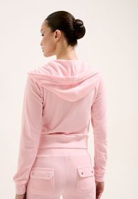 Haut à capuche rose, texture douce, coupe ajustée et manches longues. Associé à un pantalon rose assorti avec deux poches à boutons pression.