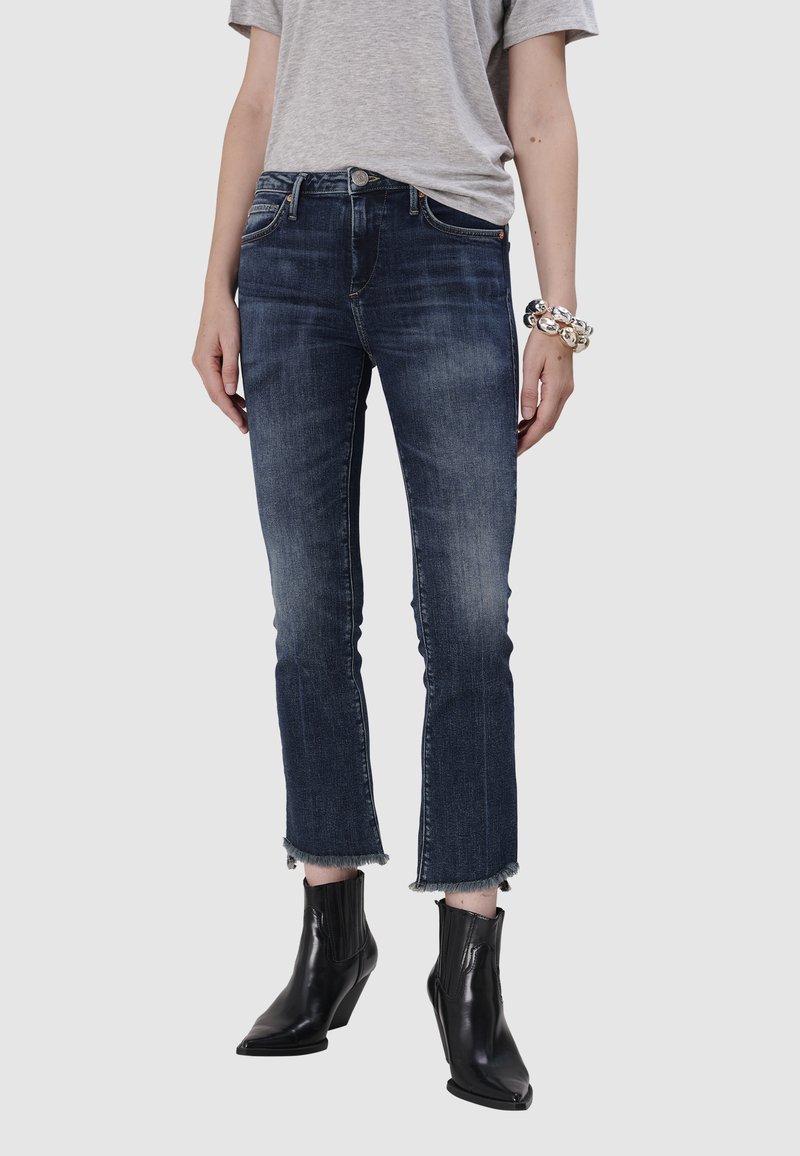 Jean en denim bleu foncé avec une finition délavée, coupé à la cheville avec des bords effilochés. Porté avec des bottines noires et un t-shirt gris.