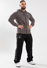 Felpa grigia con zip e maniche a costine, dettagli rossi e logo. Pantaloni della tuta neri con stampa grafica e branding bianco. Scarpe da ginnastica bianche.