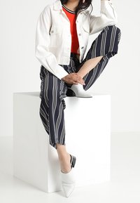 Kvinna som sitter på en vit kub iförd vit jacka, röd topp, marinblå randiga byxor och vita klackskor med spetsig tå.