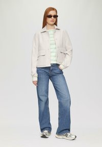 Veste courte grise clair avec de grandes poches, portée sur un haut rayé vert et blanc, associée à un jean bleu et des baskets argentées.